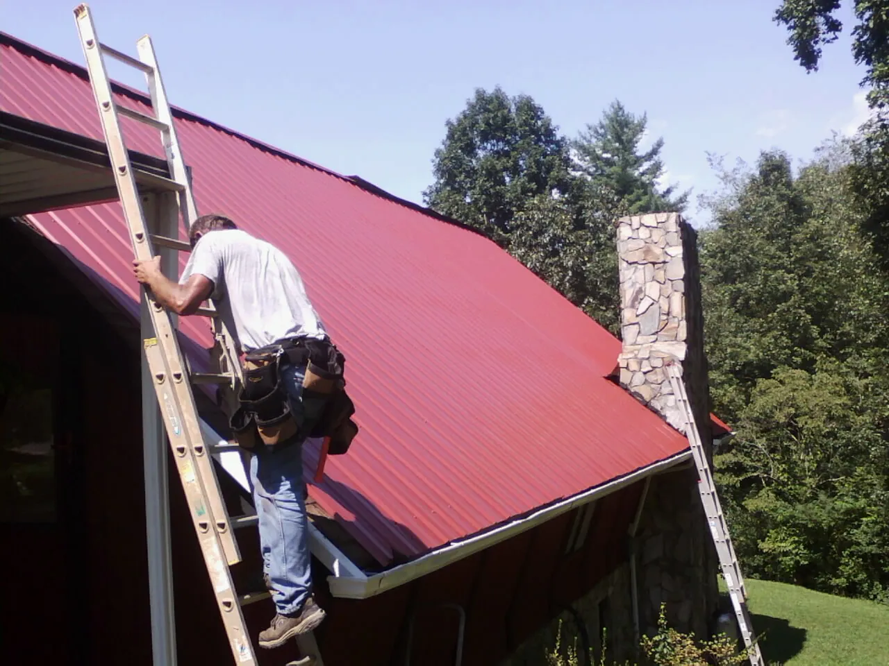 Roofer on a ladder beside a red metal roof and stone chimney — Soffit & Fascia Repair by Roofing Brothers in Berwyn